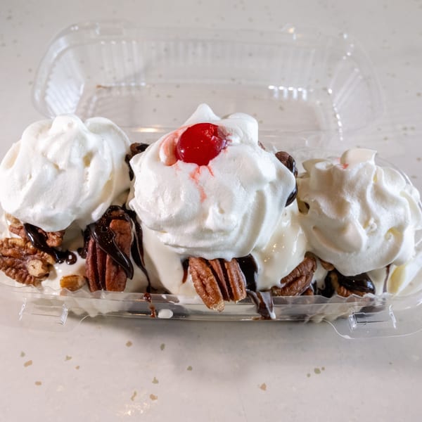 A banana split with whipped cream, chocolate syrup, pecans, and a cherry in a plastic container.
