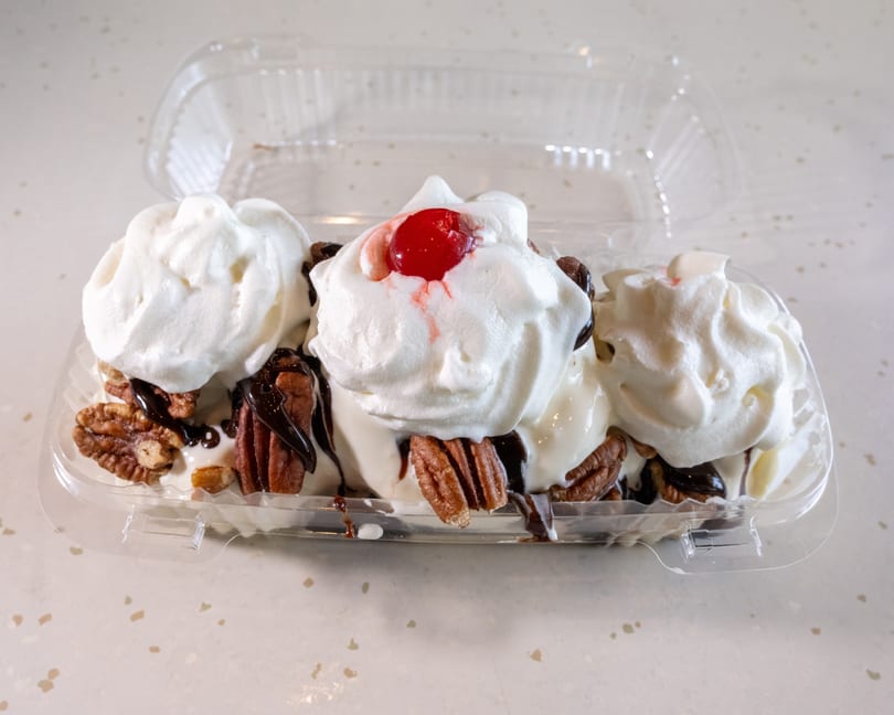 A banana split with whipped cream, chocolate syrup, pecans, and a cherry in a plastic container.