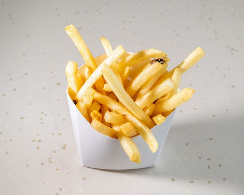 Golden french fries in a white paper container on a speckled white surface.