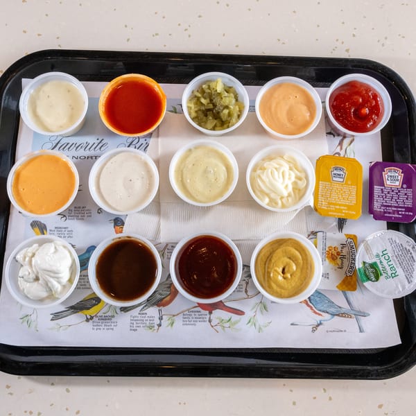 A black tray holds 15 small cups of various sauces, including ketchup, mustard, ranch, and honey.