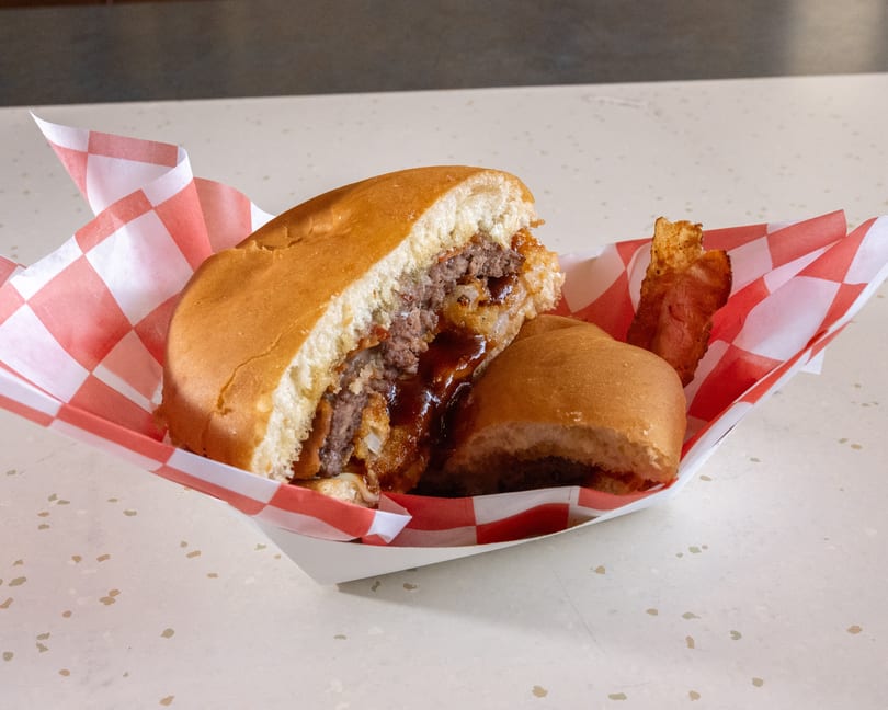A cut-in-half burger with BBQ sauce and bacon in a red and white checkered paper tray.