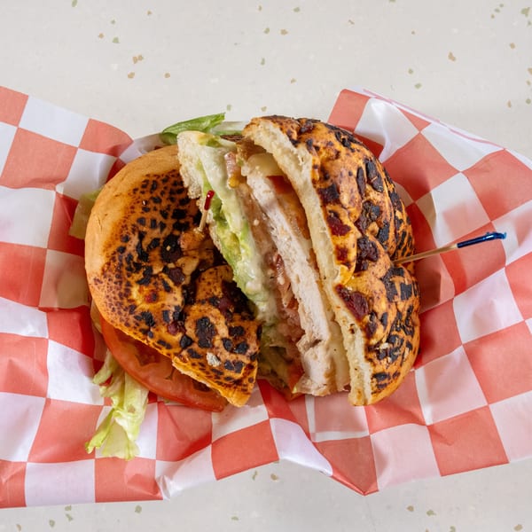 A grilled chicken sandwich, cut in half, with avocado, lettuce, tomato, and cheese on checkered paper.