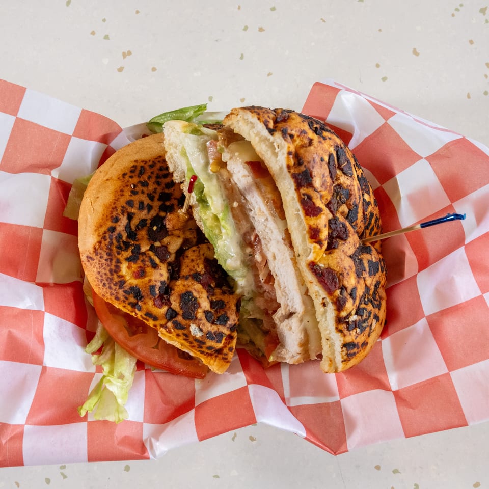 A grilled chicken sandwich, cut in half, with avocado, lettuce, tomato, and cheese on checkered paper.