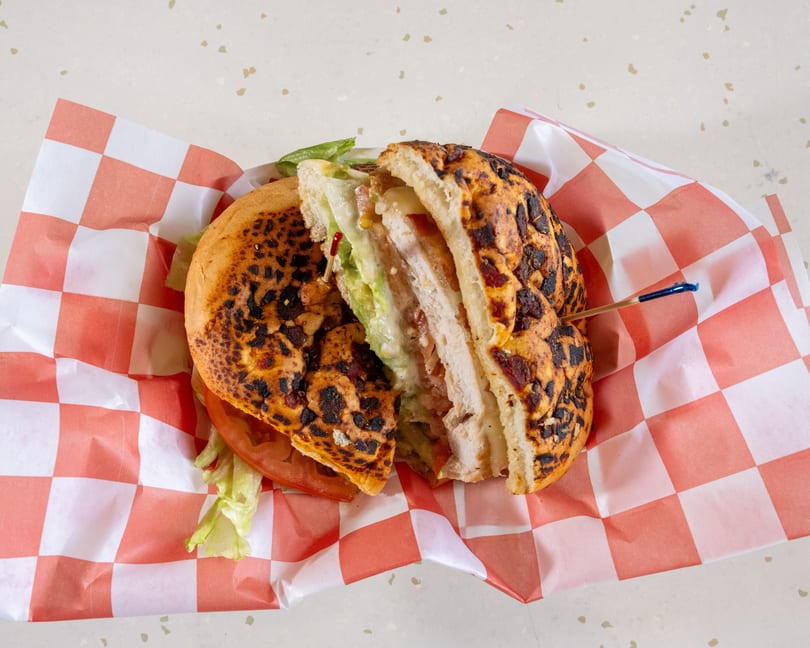 A grilled chicken sandwich, cut in half, with avocado, lettuce, tomato, and cheese on checkered paper.