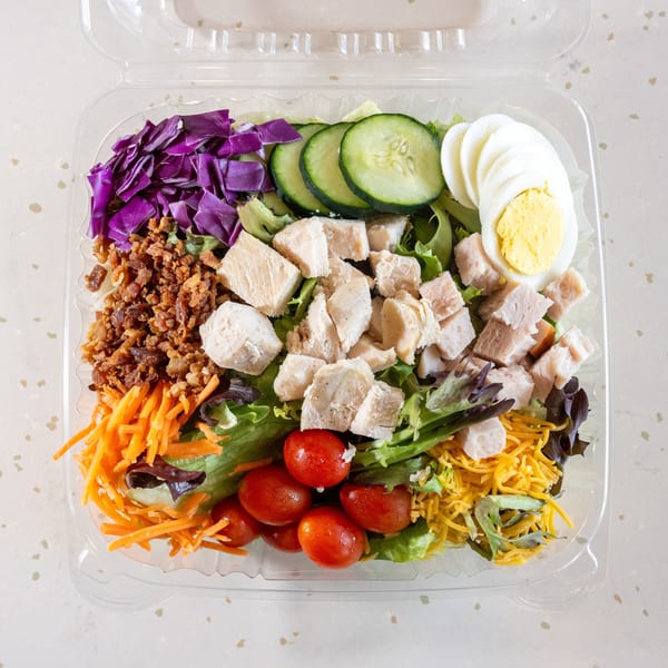 A colorful salad in a clear plastic container with chicken, egg, bacon, and vegetables.