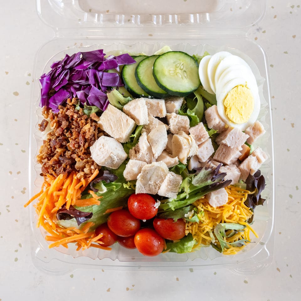 A colorful salad in a clear plastic container with chicken, egg, bacon, and vegetables.