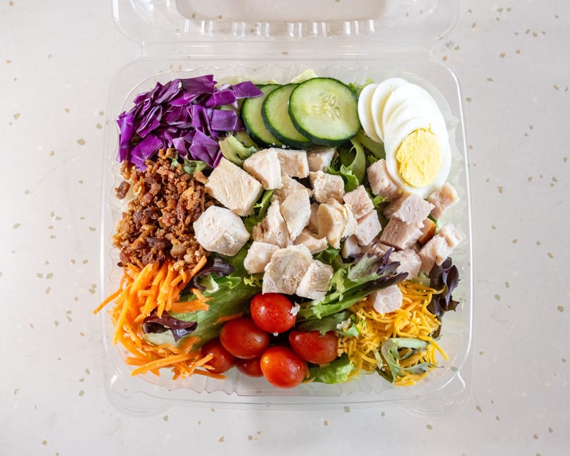 A colorful salad in a clear plastic container with chicken, egg, bacon, and vegetables.