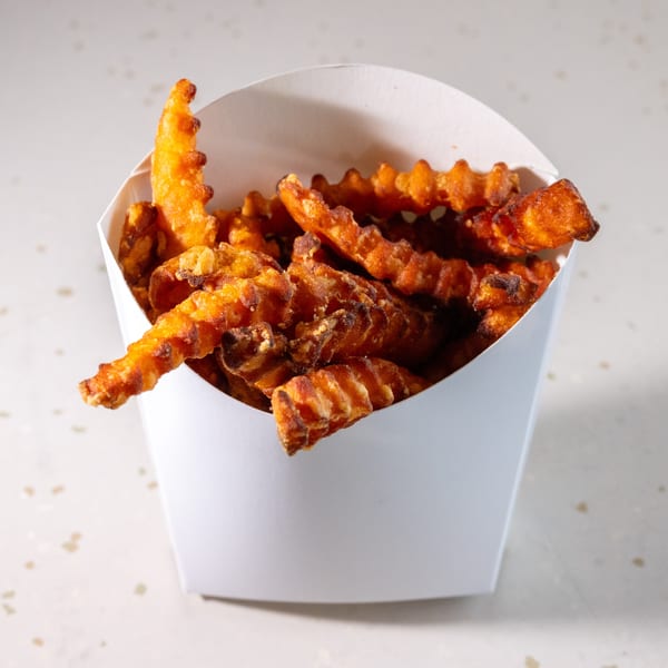 Crispy, golden-brown sweet potato waffle fries overflowing from a white paper container on a speckled surface.