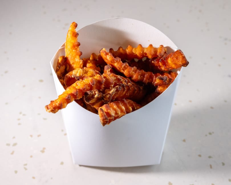 Crispy, golden-brown sweet potato waffle fries overflowing from a white paper container on a speckled surface.
