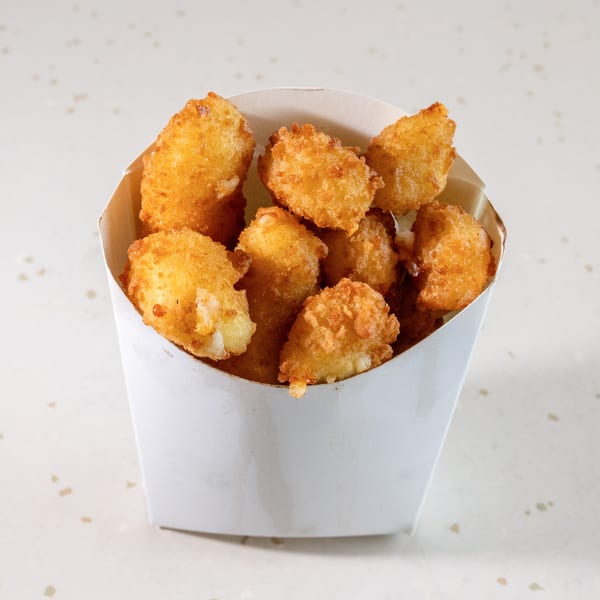 Golden-brown fried cheese curds in a white paper container on a speckled white surface.