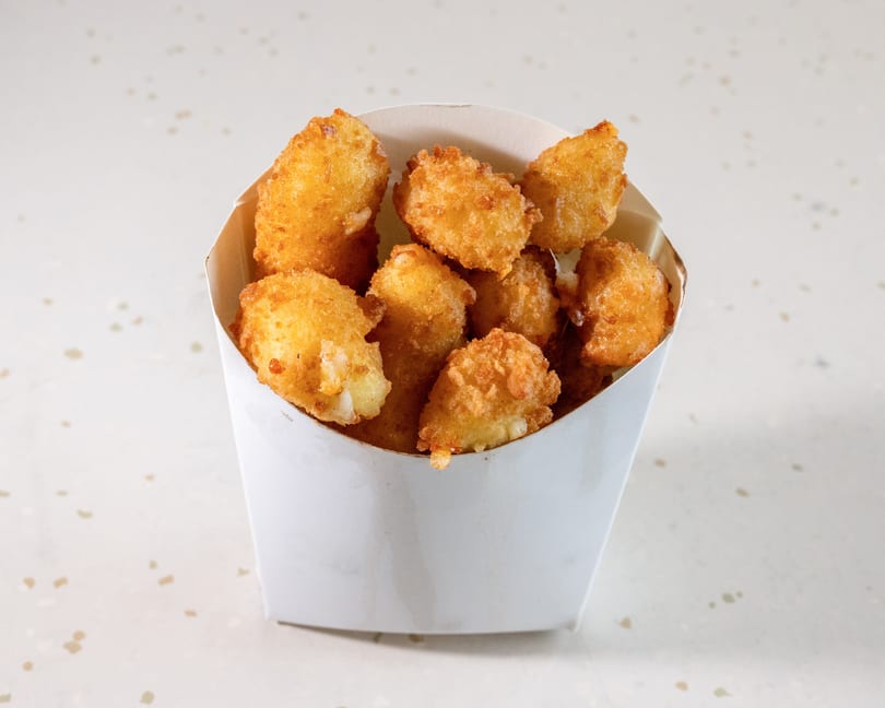 Golden-brown fried cheese curds in a white paper container on a speckled white surface.