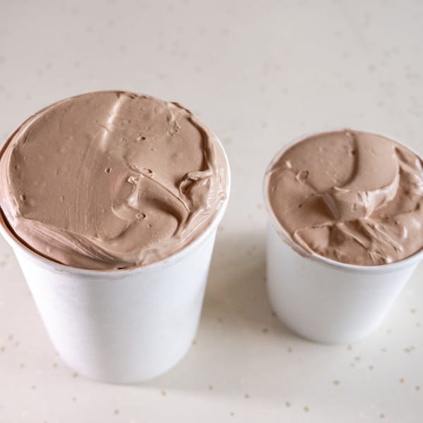 Two white cups filled with creamy chocolate ice cream sit on a light speckled surface.
