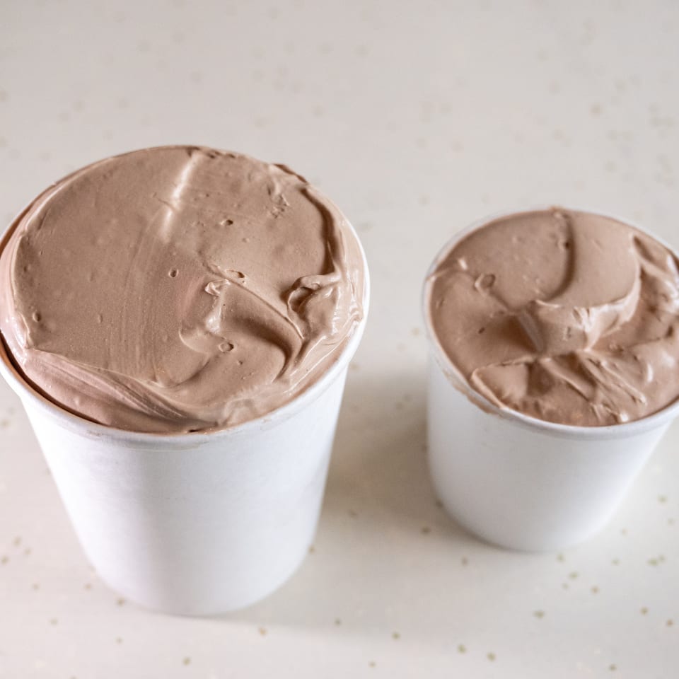 Two white cups filled with creamy chocolate ice cream sit on a light speckled surface.