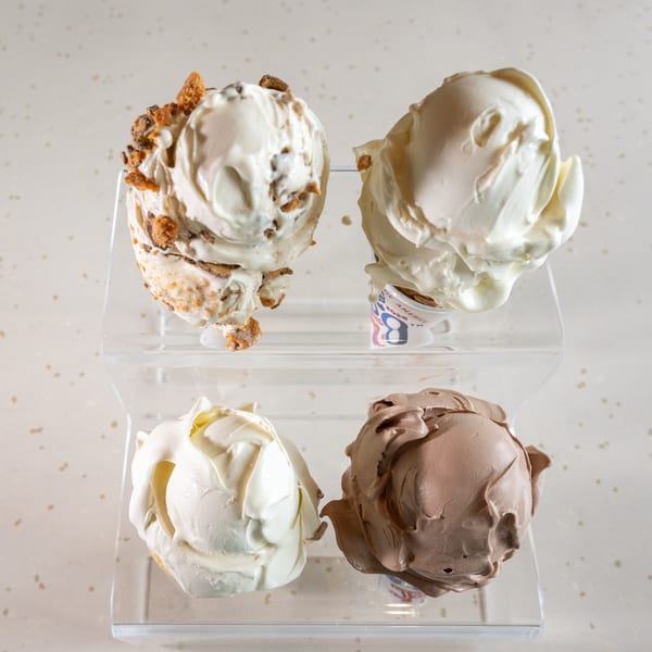 Four scoops of ice cream, two vanilla, one chocolate, and one with toppings, on a clear stand.