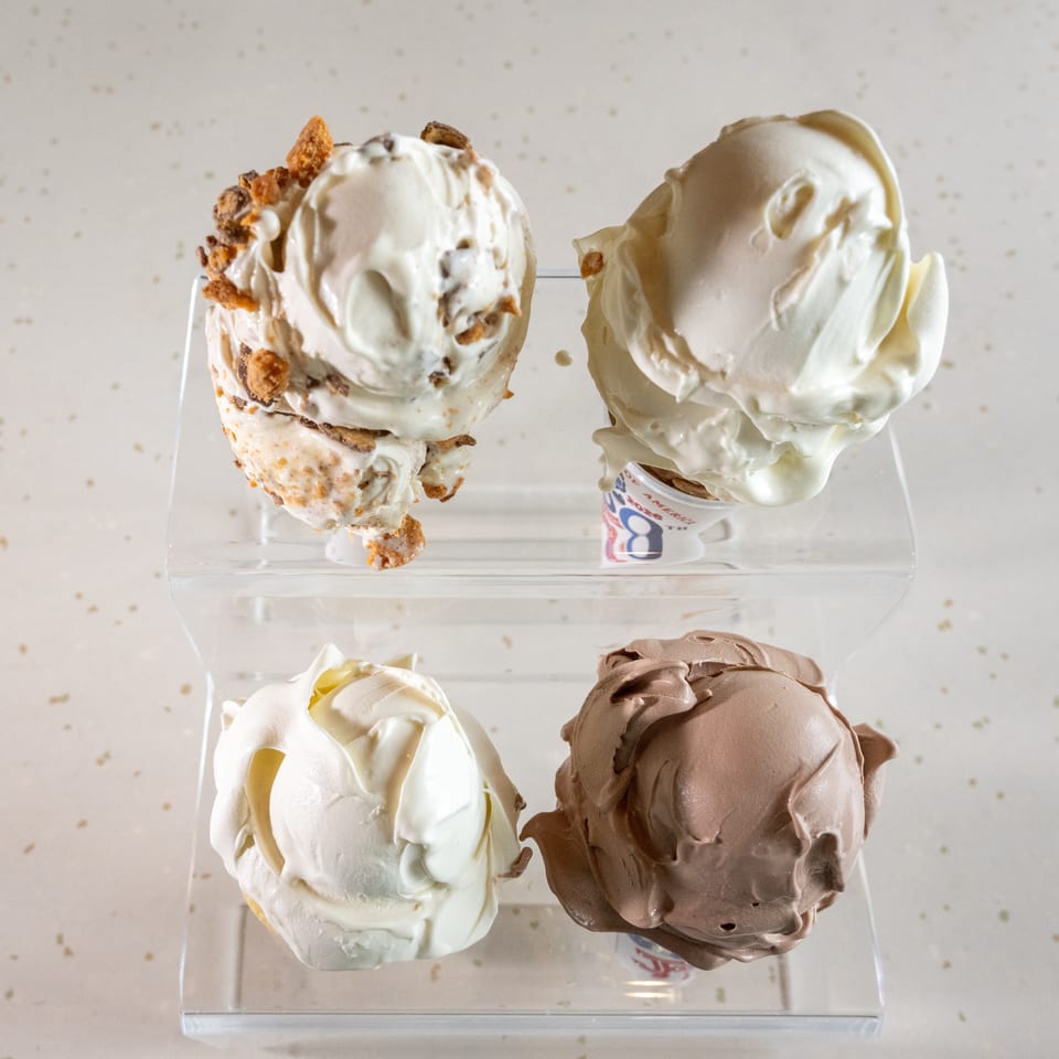 Four scoops of ice cream, two vanilla, one chocolate, and one with toppings, on a clear stand.