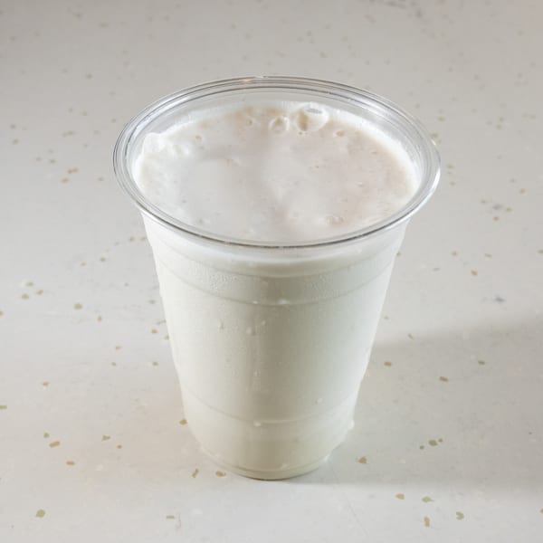 A clear plastic cup filled with a white, frothy milkshake, condensation on the outside.