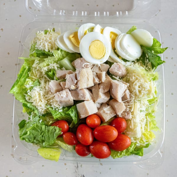 A fresh salad in a clear plastic container with lettuce, chicken, tomatoes, cheese, and hard-boiled eggs.