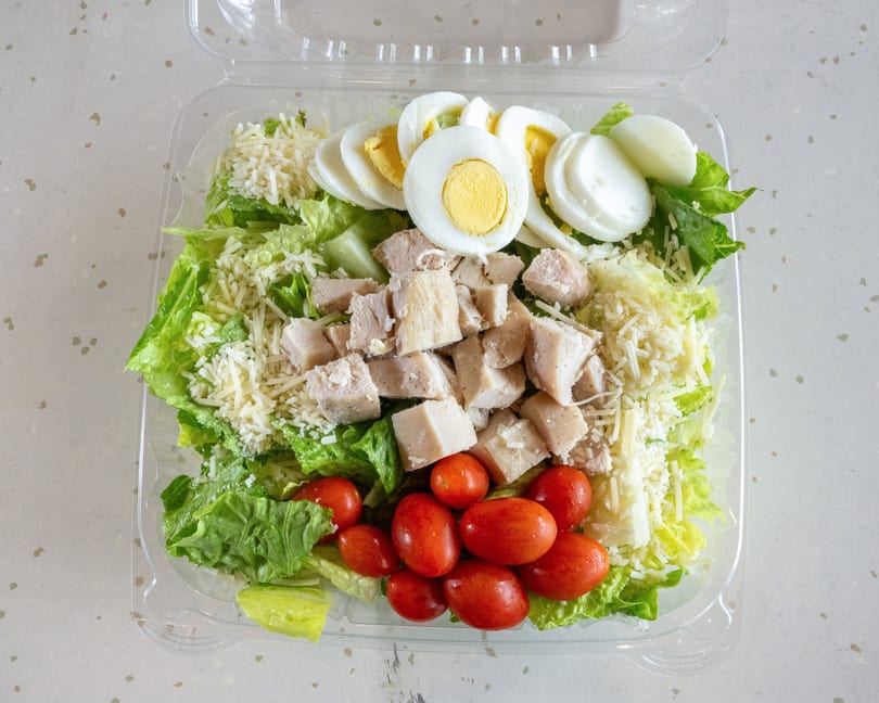 A fresh salad in a clear plastic container with lettuce, chicken, tomatoes, cheese, and hard-boiled eggs.